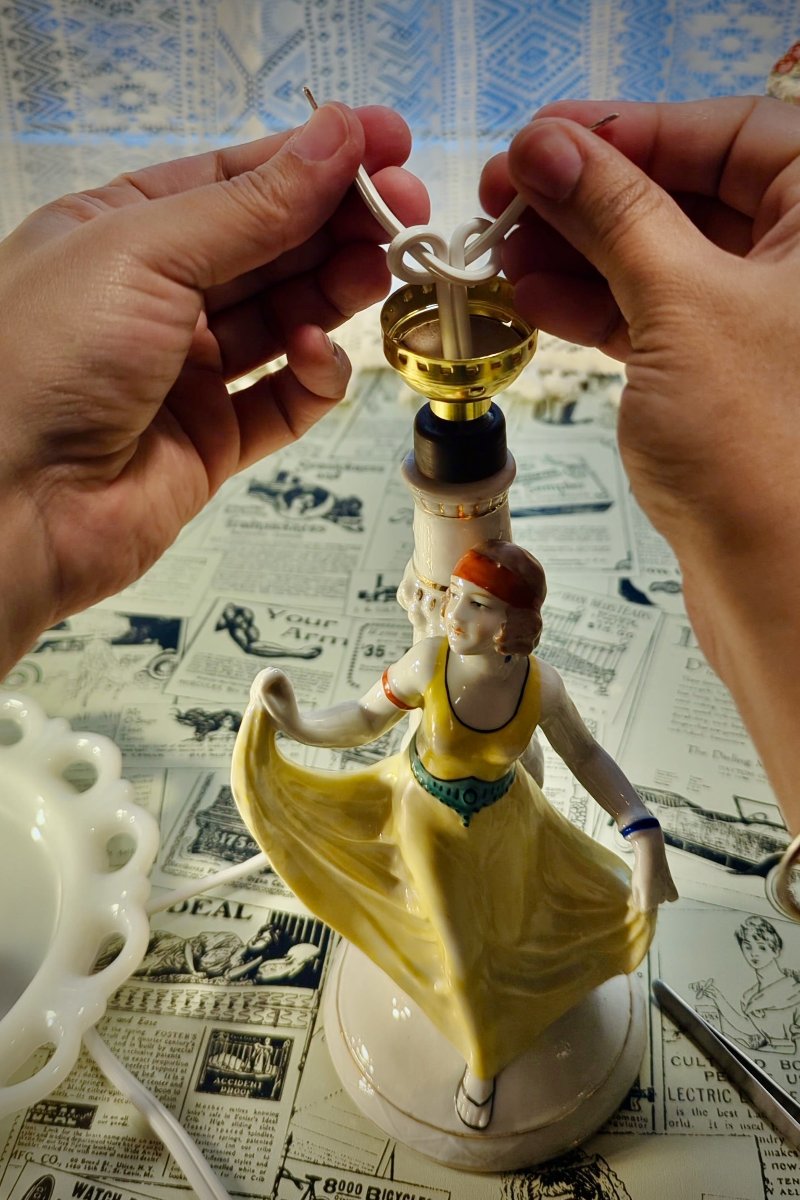 The artist is tying an underwriters knot to secure electrical wires onto a ceramic figural lampshade of a woman holding the hem of her yellow dress. 